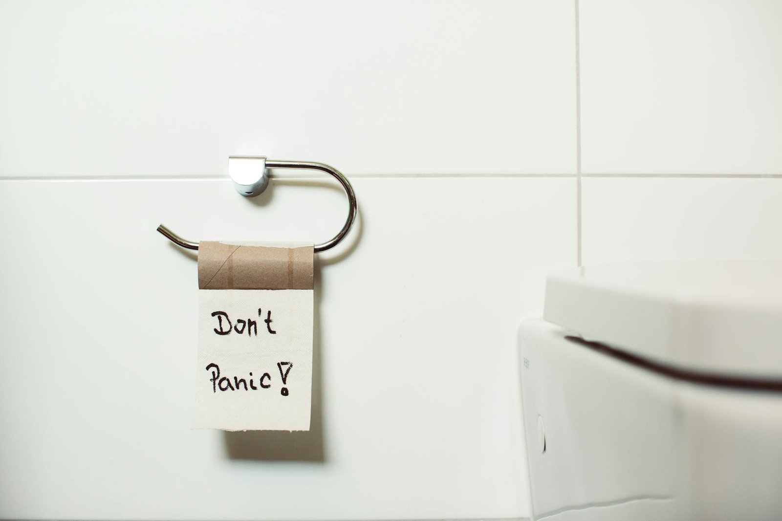 A toilet paper holder with an empty roll displaying a 'Don't Panic!' note, set in a modern bathroom.
