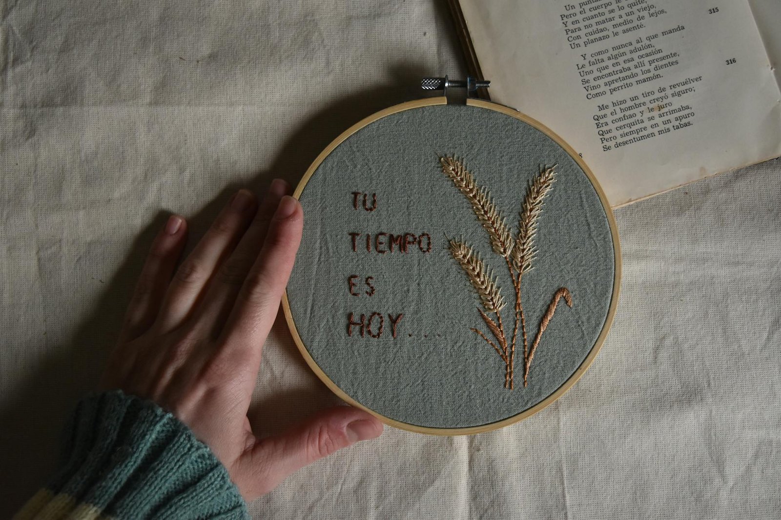 A hand touches an embroidery hoop with Spanish text and flowers beside an open book.
