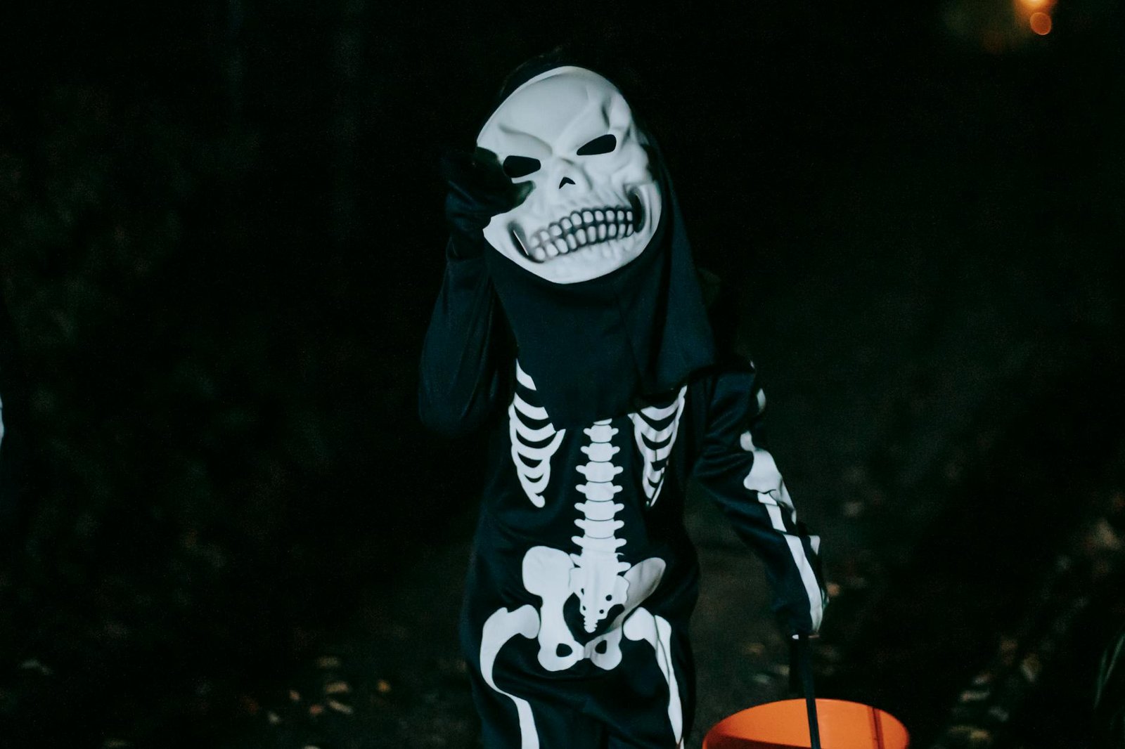 A child dressed in a skeleton costume with a bucket, trick-or-treating on Halloween night outdoors.