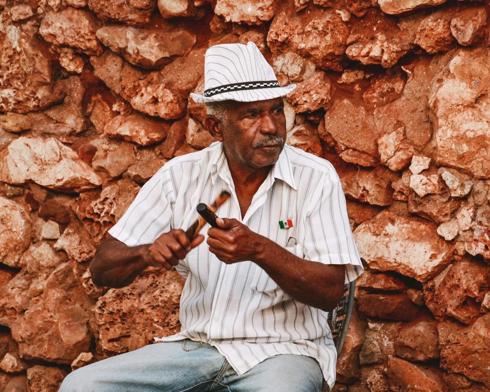 A senior musician plays claves against a rustic stone wall, adorned with a Mexican flag pin.