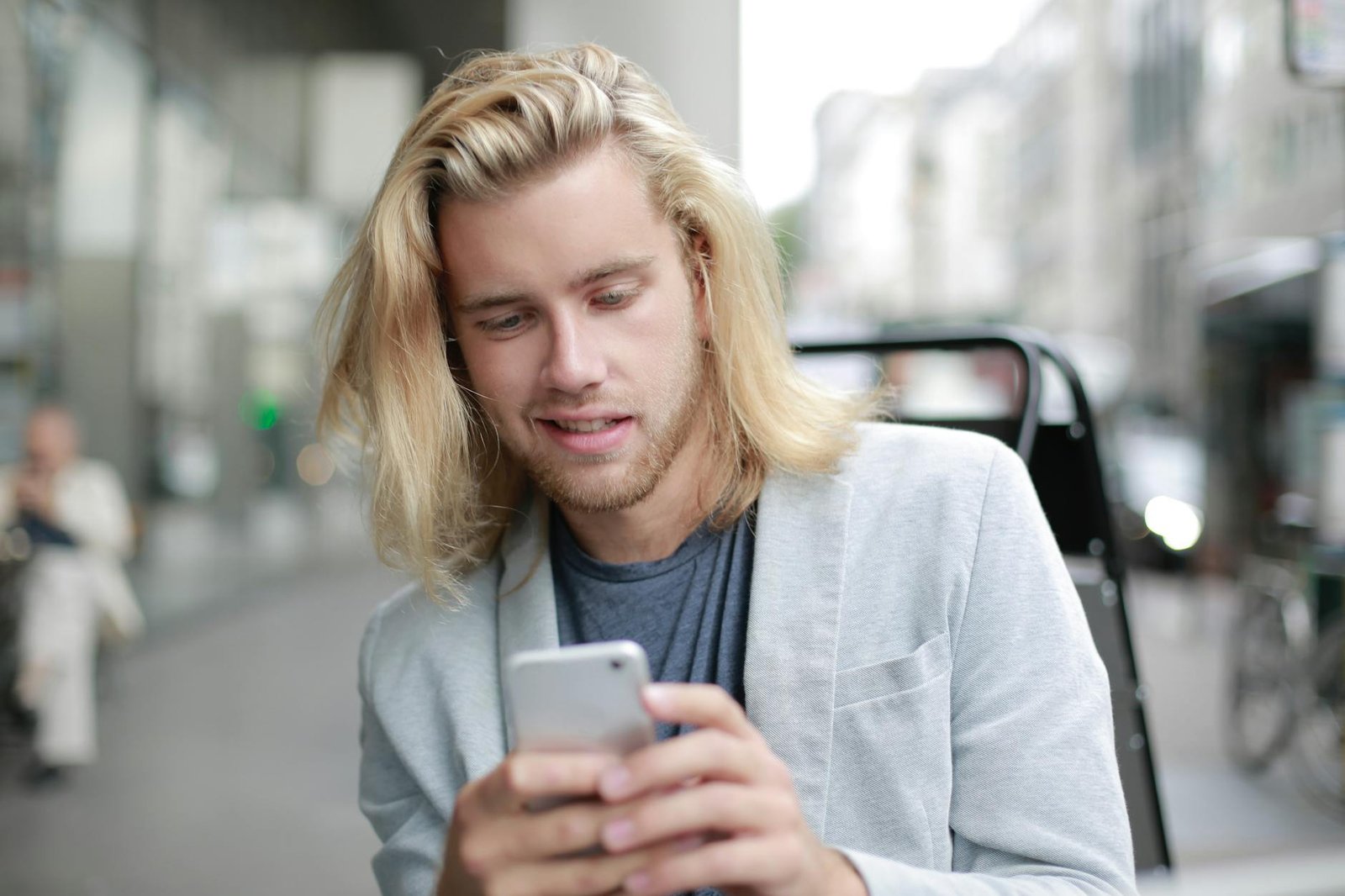 Smiling young man with long blonde hair using a smartphone while outdoors in an urban setting.