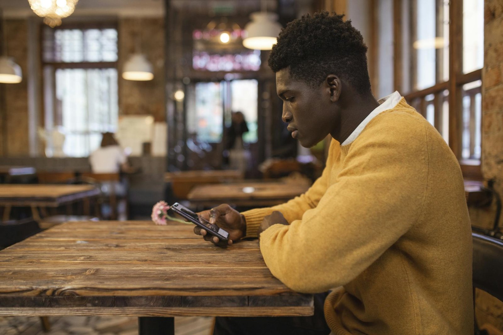 A man in a yellow sweater focused on his smartphone while sitting in a rustic café.