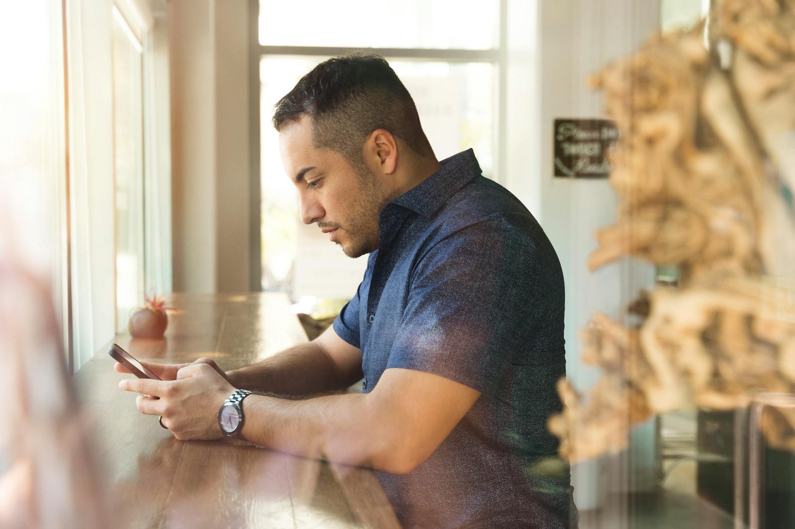 Man in a blue shirt using smartphone indoors by a window at a café.