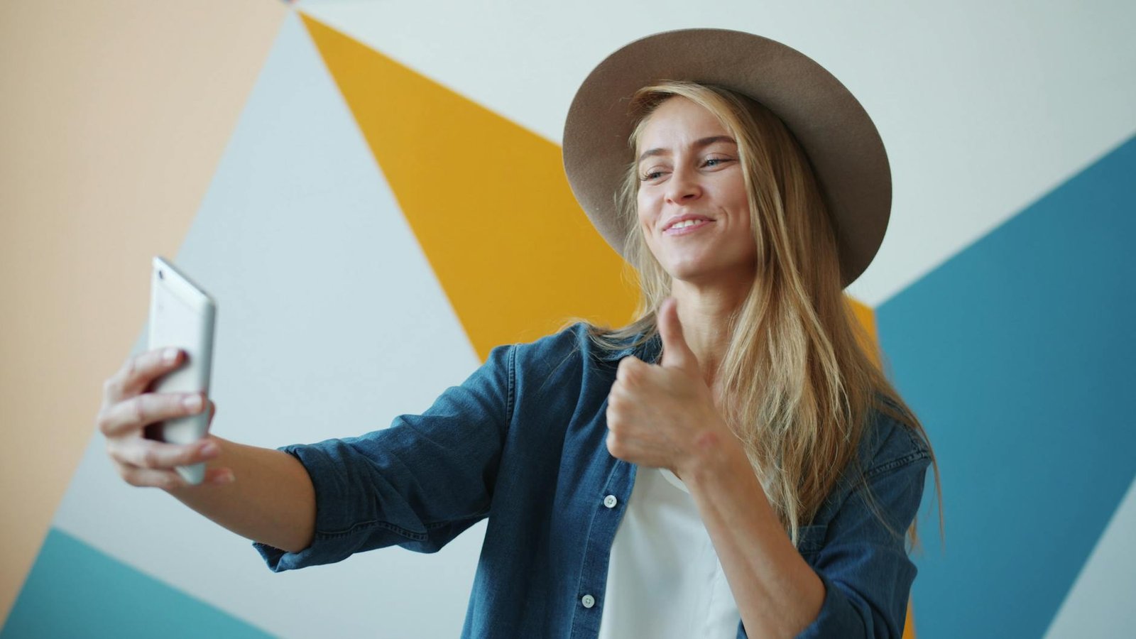 Happy woman wearing hat taking a selfie with thumbs up in front of geometric background.