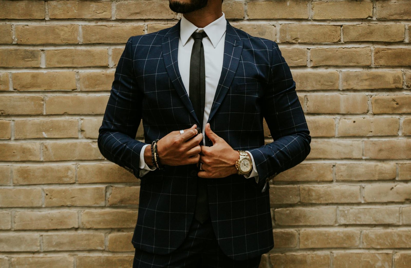 Elegant businessman adjusts his checkered suit and tie against a brick wall.