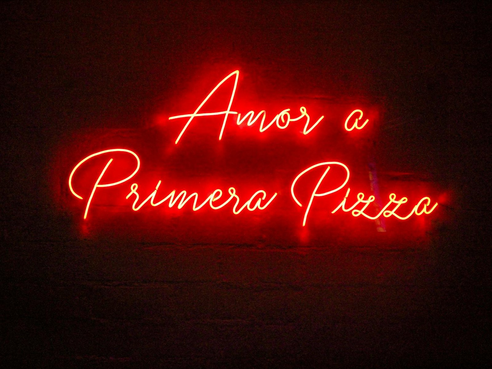 Vibrant red neon sign displaying 'Amor a Primera Pizza' on a dark background, adding a vintage, romantic touch.