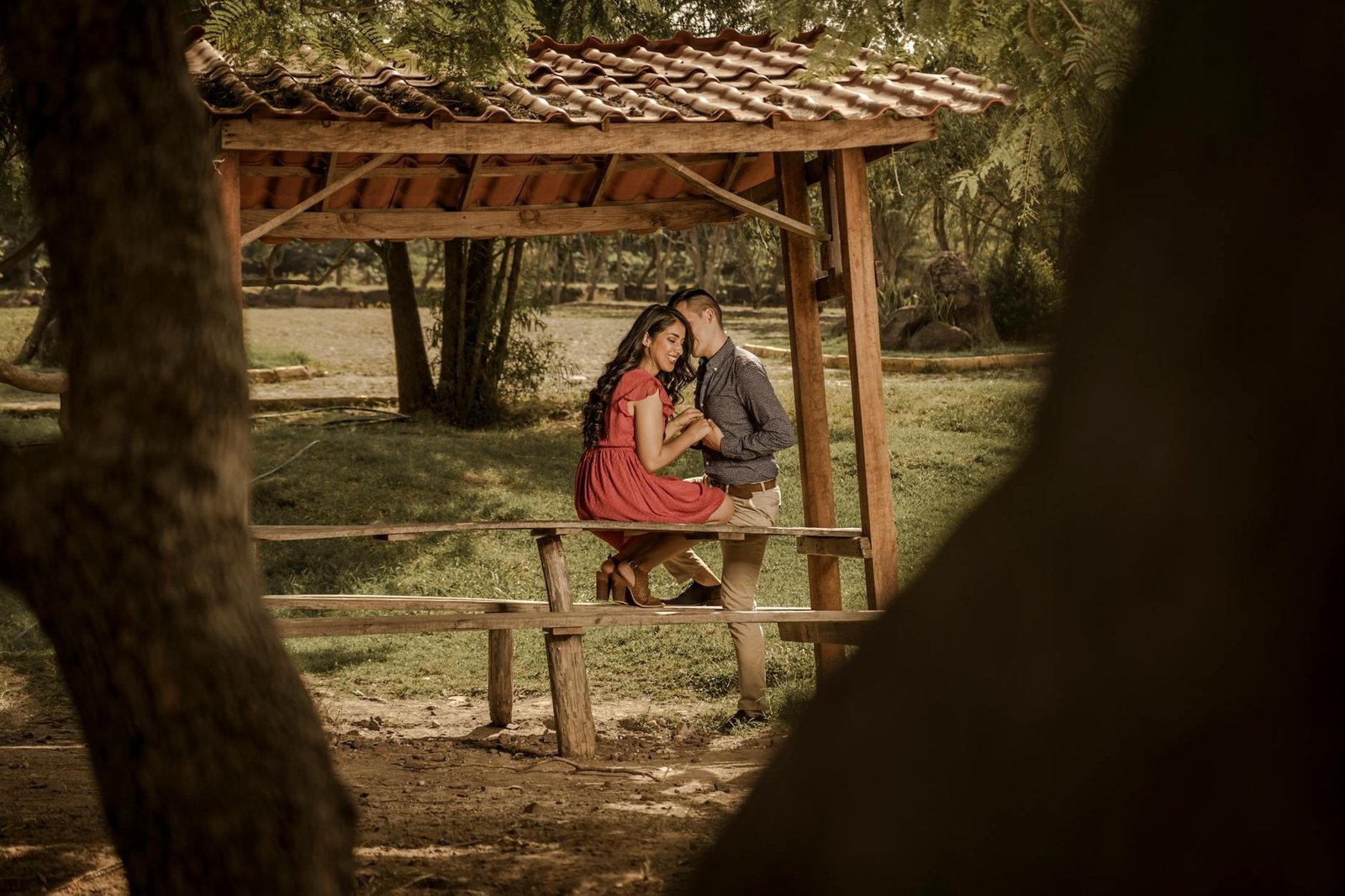 A loving couple sitting on a picnic table, sharing a tender moment under a roofed structure in a serene outdoor setting.