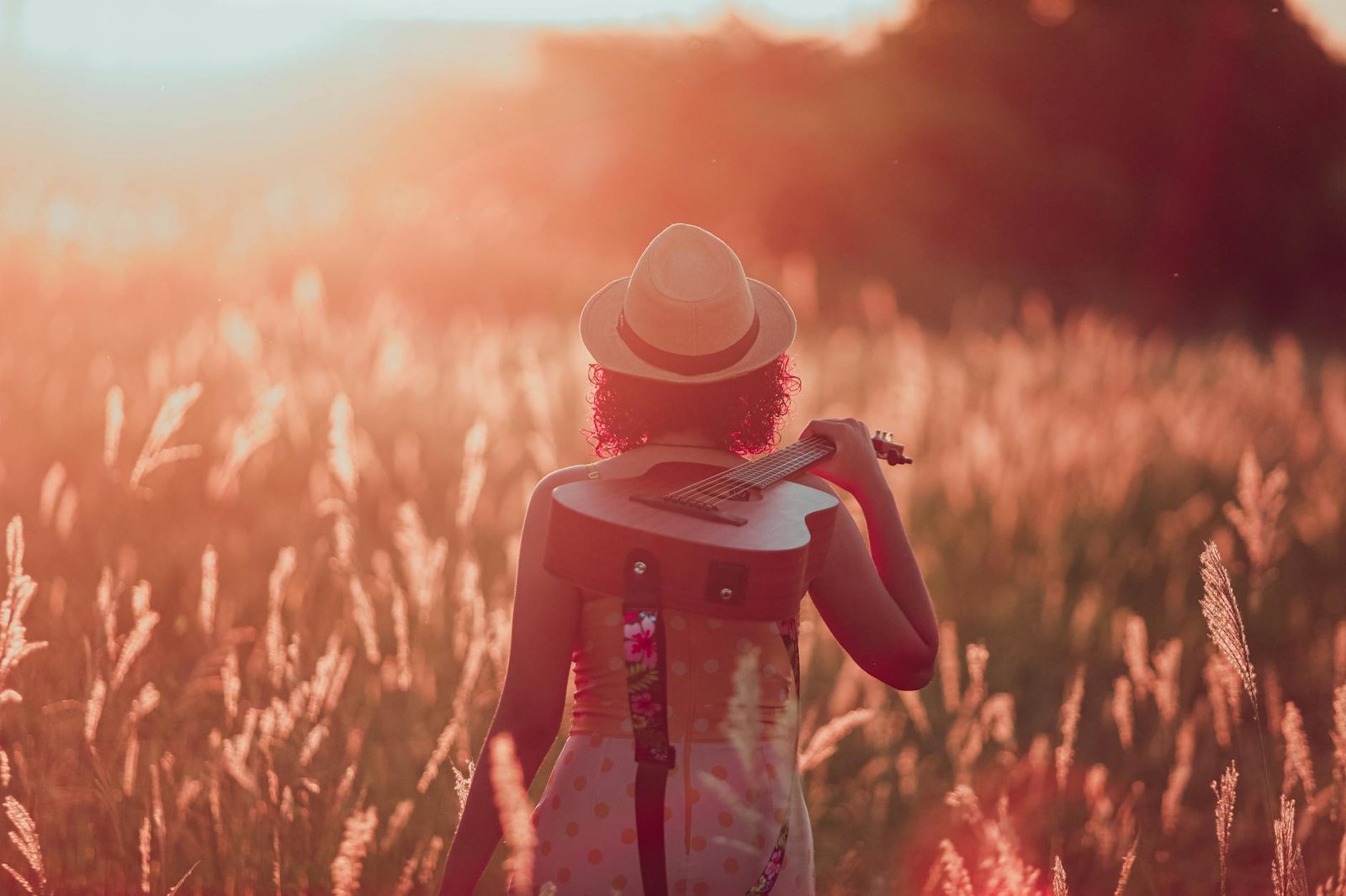 A woman in a hat carrying a guitar through a sunlit field at sunset in Paragominas, Brazil.
