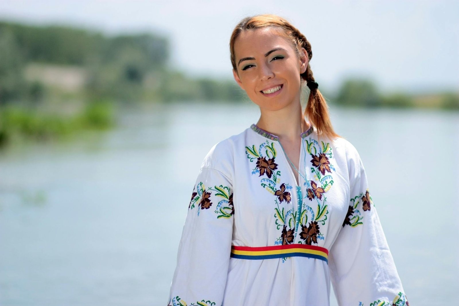 A cheerful woman in traditional embroidered clothing posing by a scenic river outdoors.