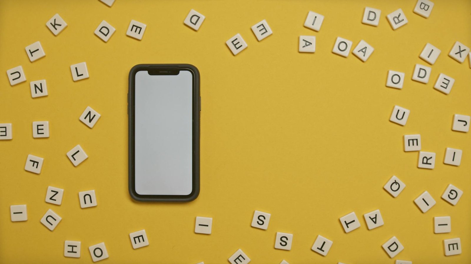 Smartphone surrounded by scattered alphabet tiles on a vibrant yellow background.